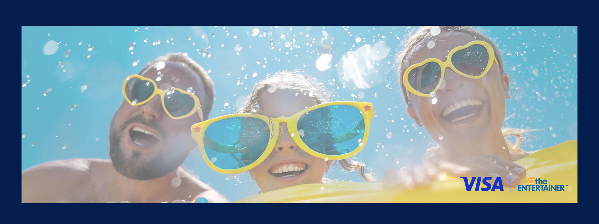 Three people smiling underwater, wearing oversized yellow sunglasses. Bubbles surround them in the bright blue water. Logos of Visa and The Entertainer are visible.