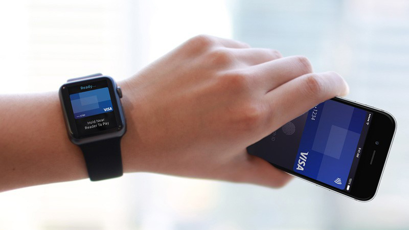 A left hand holding a mobile phone with a Visa card visible on its screen and an Apple watch on the wrist.