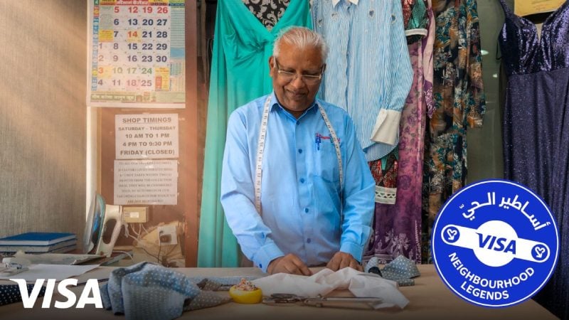 Elderly tailor smiling at his workbench, surrounded by colorful dresses. Sewing tools and a VISA Neighbourhood Legends badge are visible. 