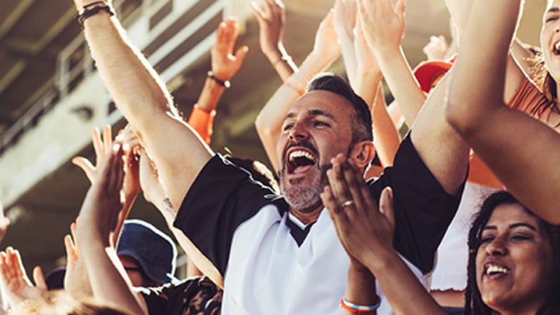 Excited crowd at a sports event. A man with raised arms cheers energetically, surrounded by joyful spectators under sunlit bleachers. 