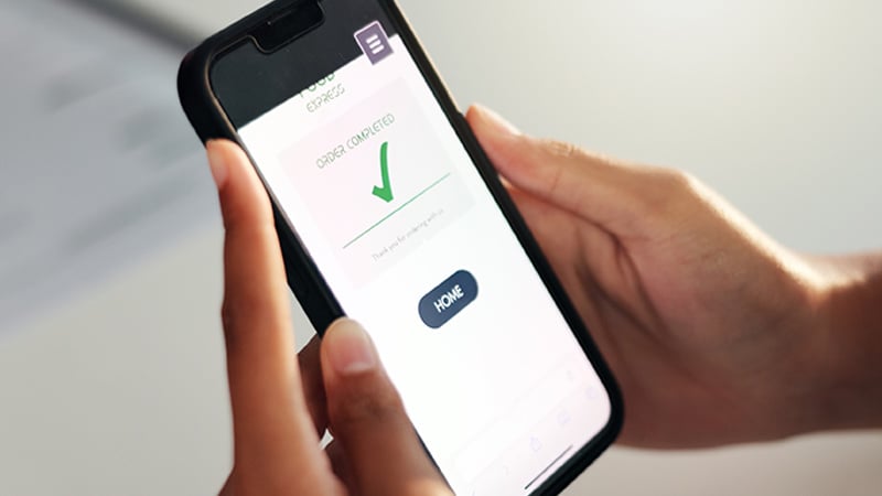 A person holding a smartphone showing an "Order Completed" screen with a green checkmark and a "Home" button, conveying a sense of achievement.