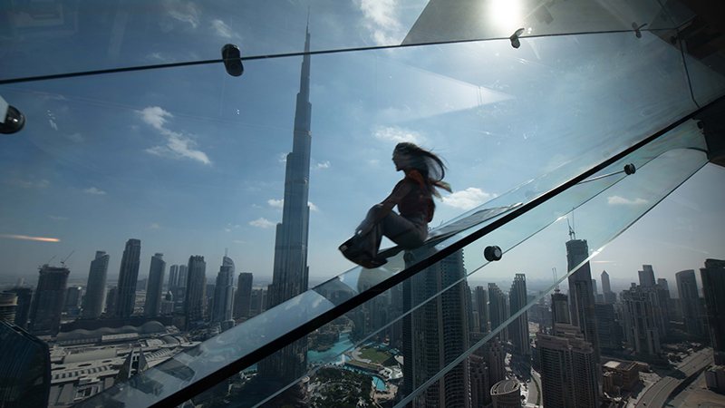 A person slides down a glass structure high above a city skyline, with the Burj Khalifa visible