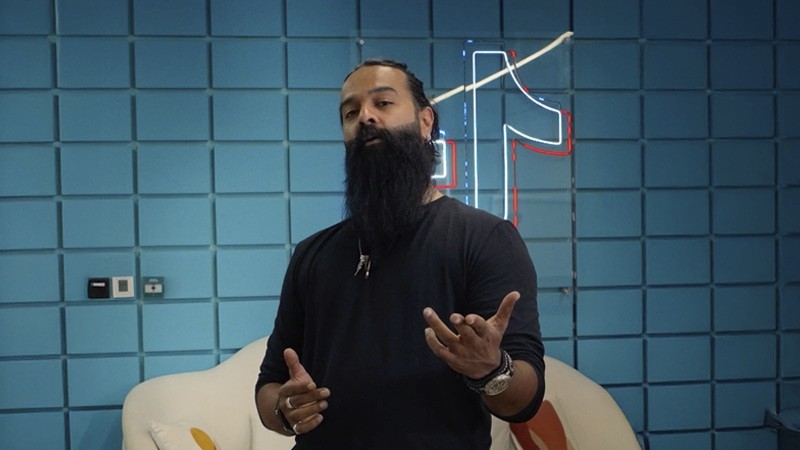 Bearded man speaking in front of a blue tiled wall with a neon music note