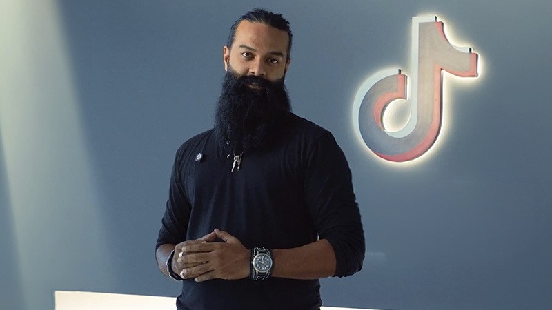 Bearded man standing beside a glowing TikTok logo on a gray wall